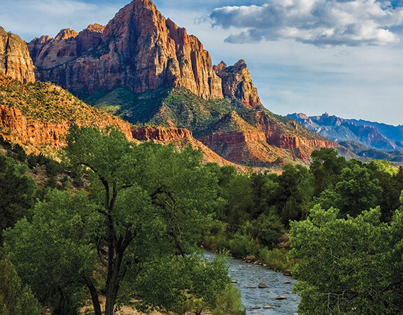 Scenic view of towering red rock mountains and lush green landscape with a river under a partly cloudy sky.