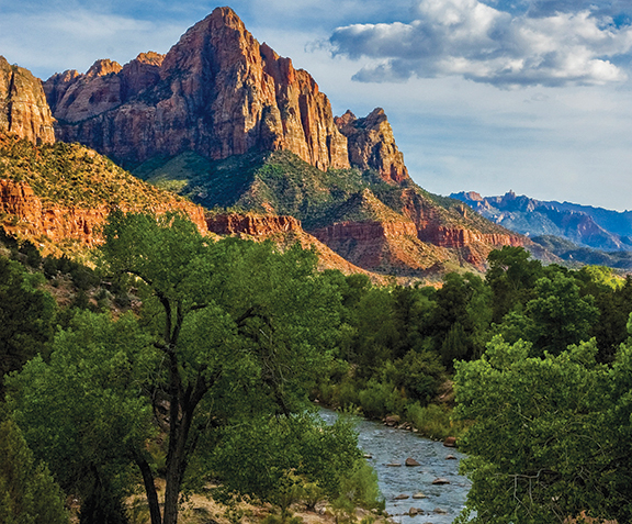 Scenic view of towering red rock mountains and lush green landscape with a river under a partly cloudy sky.