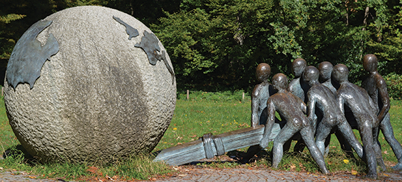 Bronze sculpture of figures pushing a large stone sphere in a park setting.