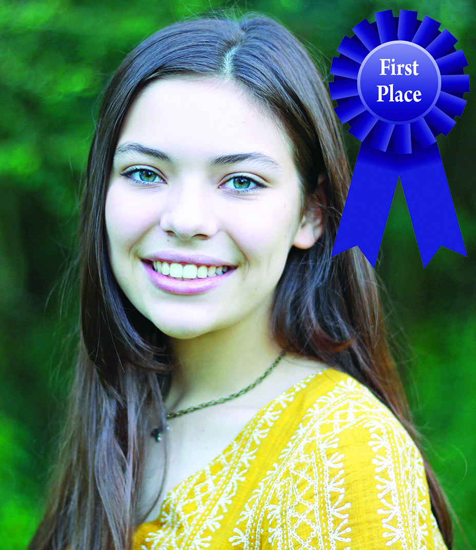 Smiling woman with long hair, wearing a yellow top, a blue First Place ribbon beside her, green background.