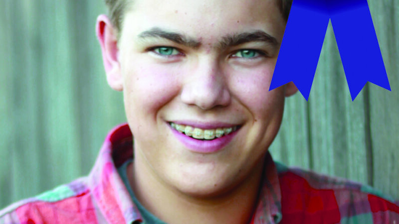 Smiling boy with braces wearing a plaid shirt, awarded a blue first-place ribbon.