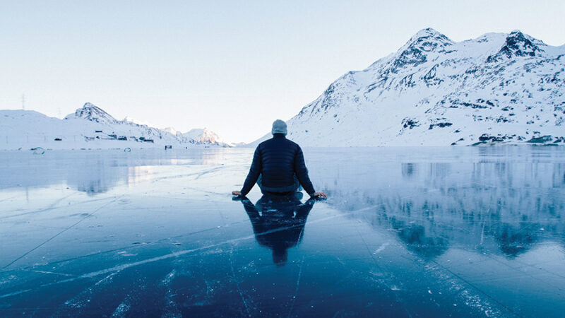 Person sitting on a frozen lake, surrounded by snowy mountains under a clear sky. Serene winter landscape.