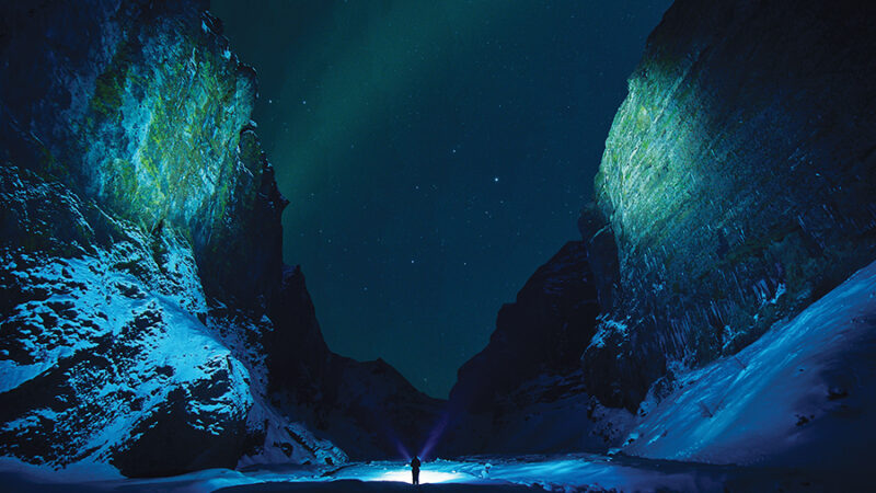 Person with flashlight in a snowy canyon under a starlit sky with green aurora borealis.