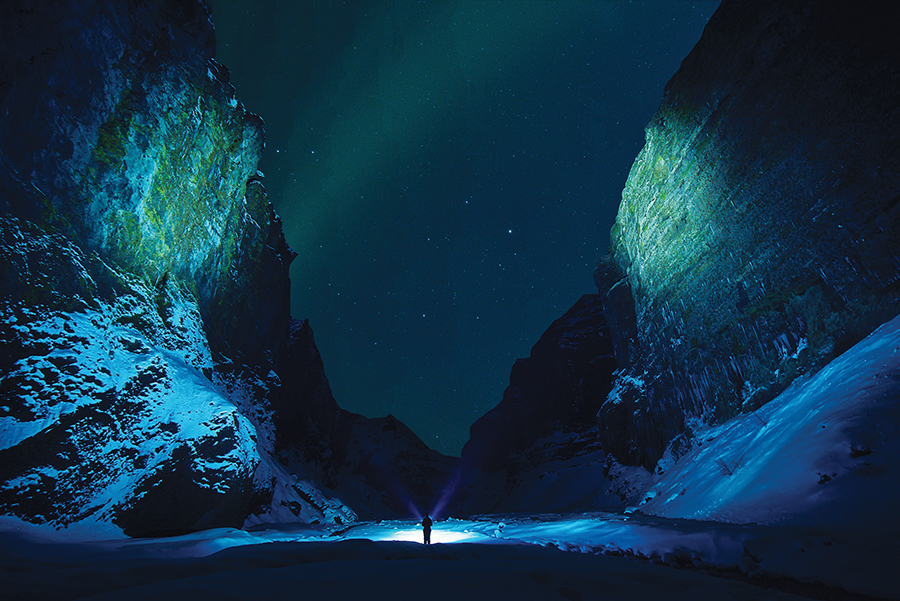 Person with flashlight in a snowy canyon under a starlit sky with green aurora borealis.