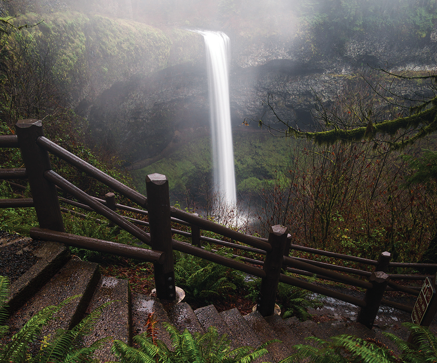 Misty waterfall view from wooden stairway in dense forest setting, emphasizing the natural beauty and tranquility of the scene.