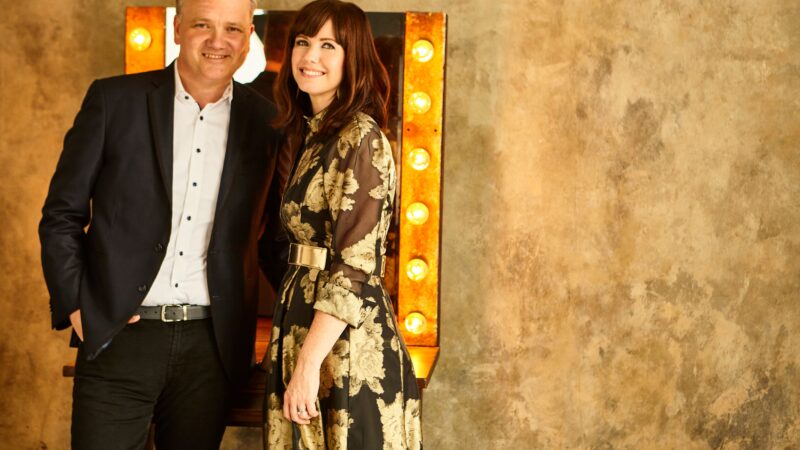 Smiling couple standing in front of rustic wall with marquee lights.