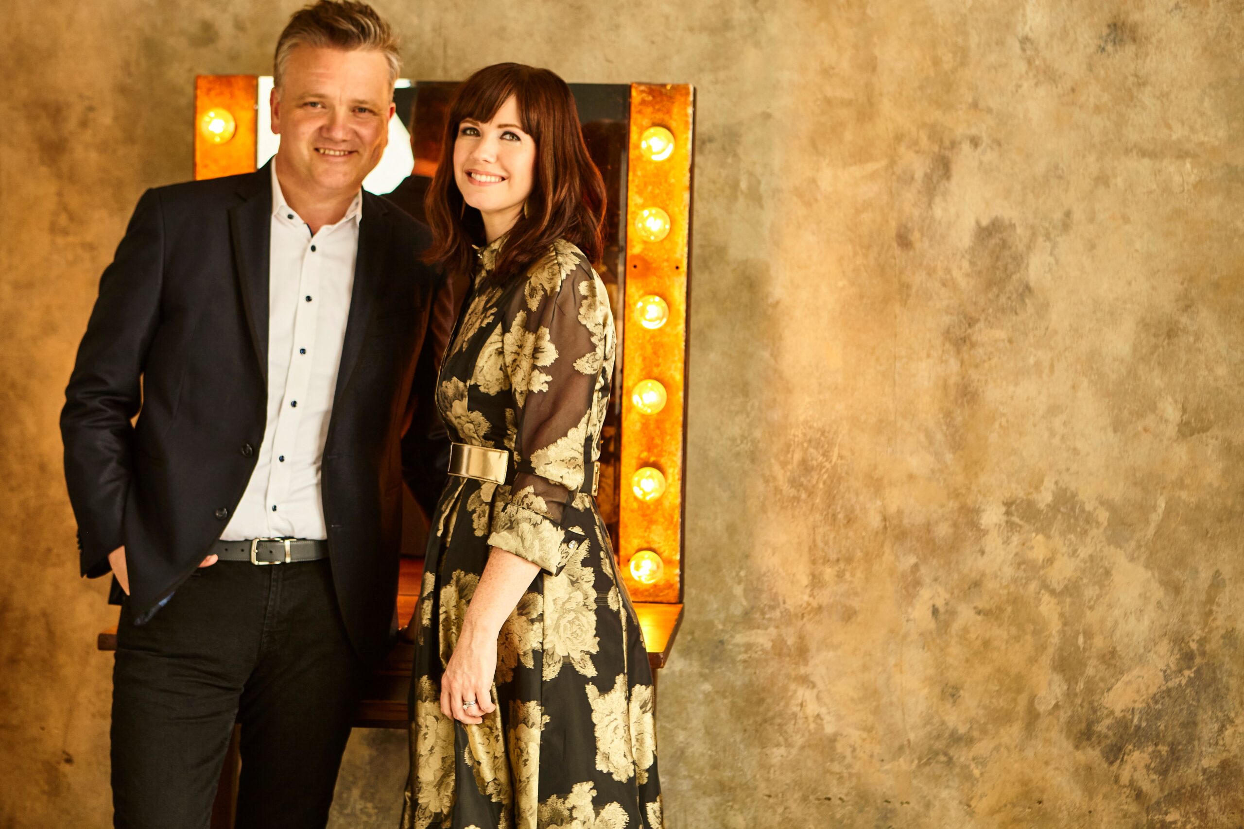 Smiling couple standing in front of rustic wall with marquee lights.