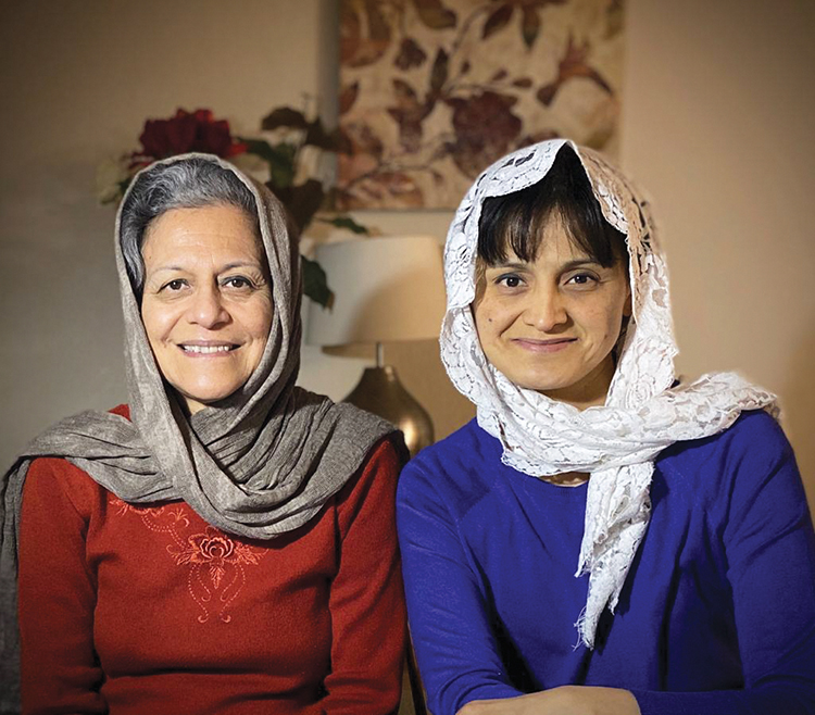 Two women smiling, wearing headscarves, sitting indoors with a warm background setting.