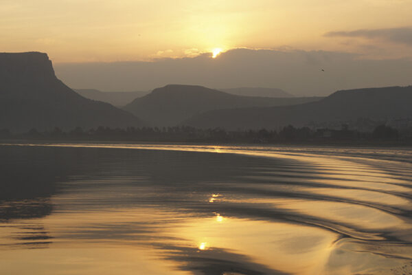 Sunset over calm water with ripples and silhouetted mountains in the background.