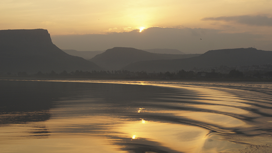 Sunset over calm water with ripples and silhouetted mountains in the background.