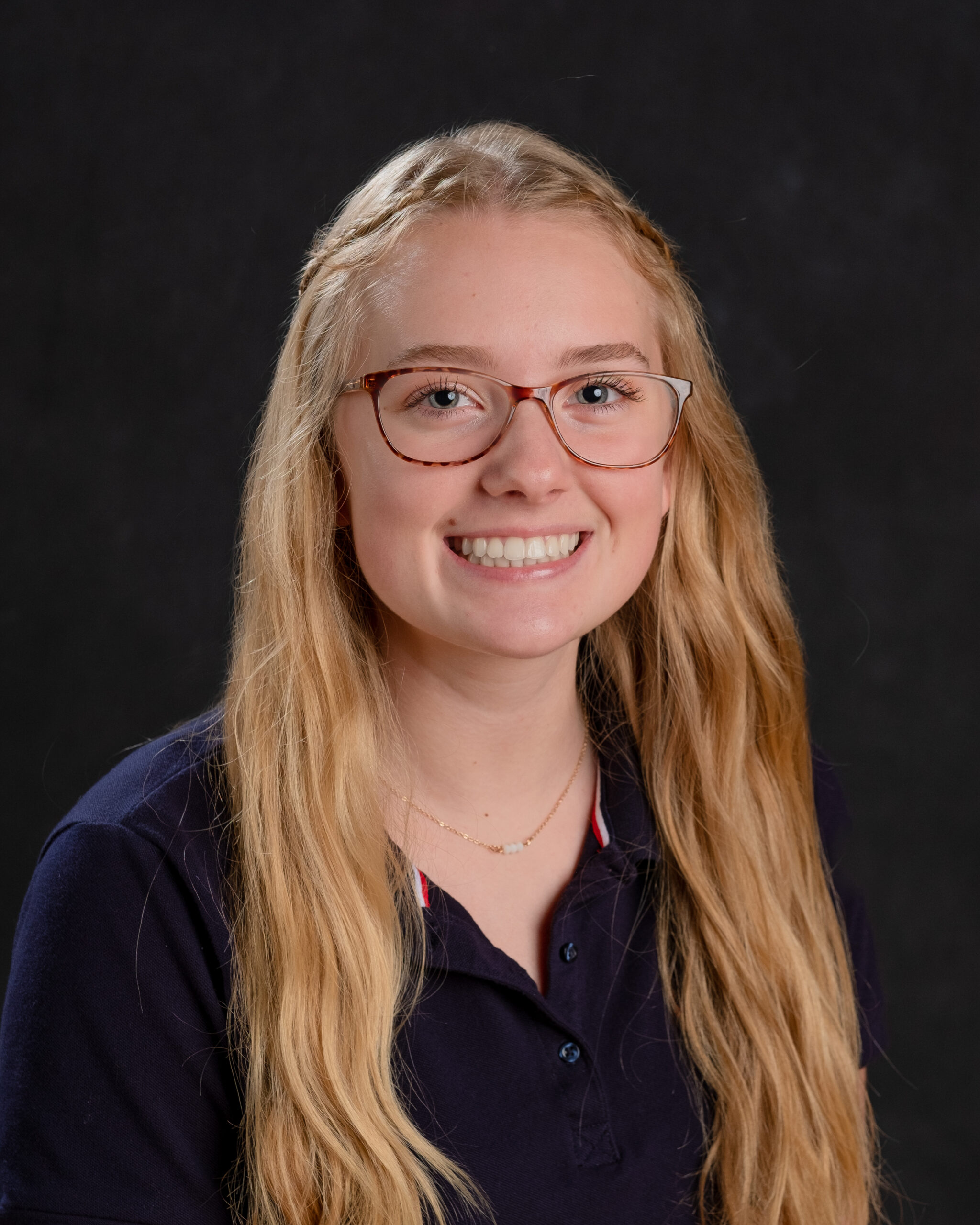 Young woman smiling with long blonde hair and glasses, wearing a dark shirt against a dark background.