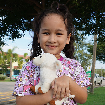 Smiling child holding a stuffed animal outdoors in a park, wearing a patterned shirt.