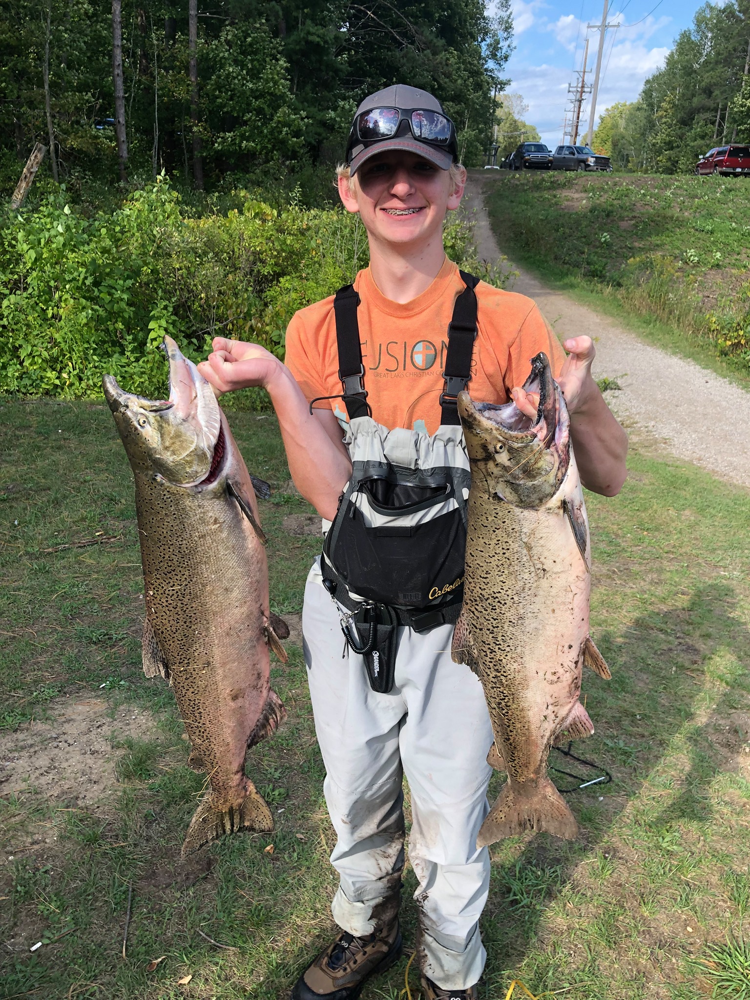 Man smiling and holding two large fish outdoors on a sunny day, with trees and a dirt path in the background.