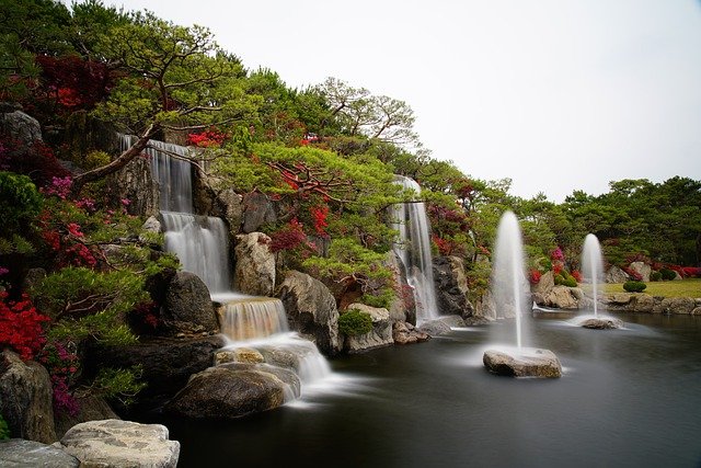 Tranquil garden scene with cascading waterfalls and fountains surrounded by vibrant greenery and rocks.