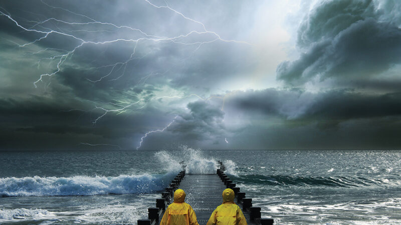 Two people in yellow raincoats hold hands on a pier during a dramatic lightning storm over the ocean.