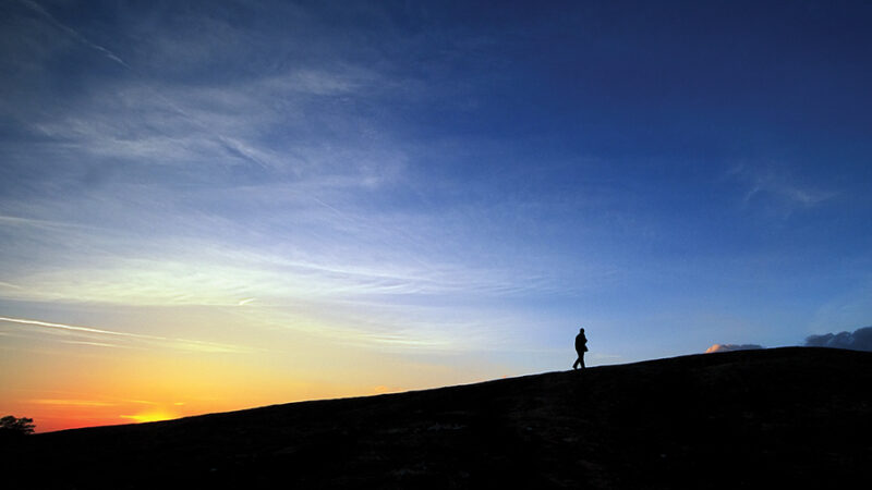 Silhouette of a person walking on a hill at sunrise, with a colorful sky and clouds in the background.