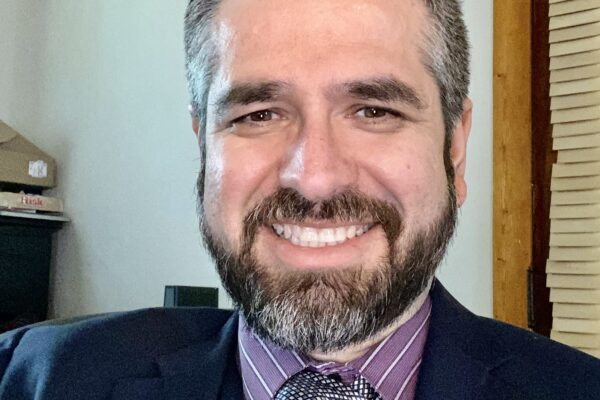 Smiling man in formal attire with beard and tie, sitting indoors near window blinds.