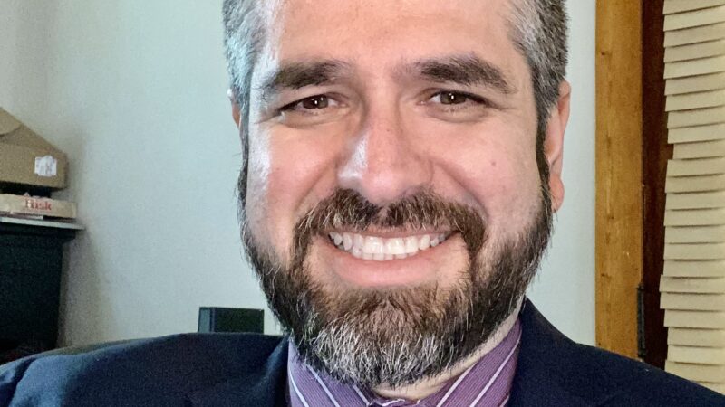 Smiling man in formal attire with beard and tie, sitting indoors near window blinds.