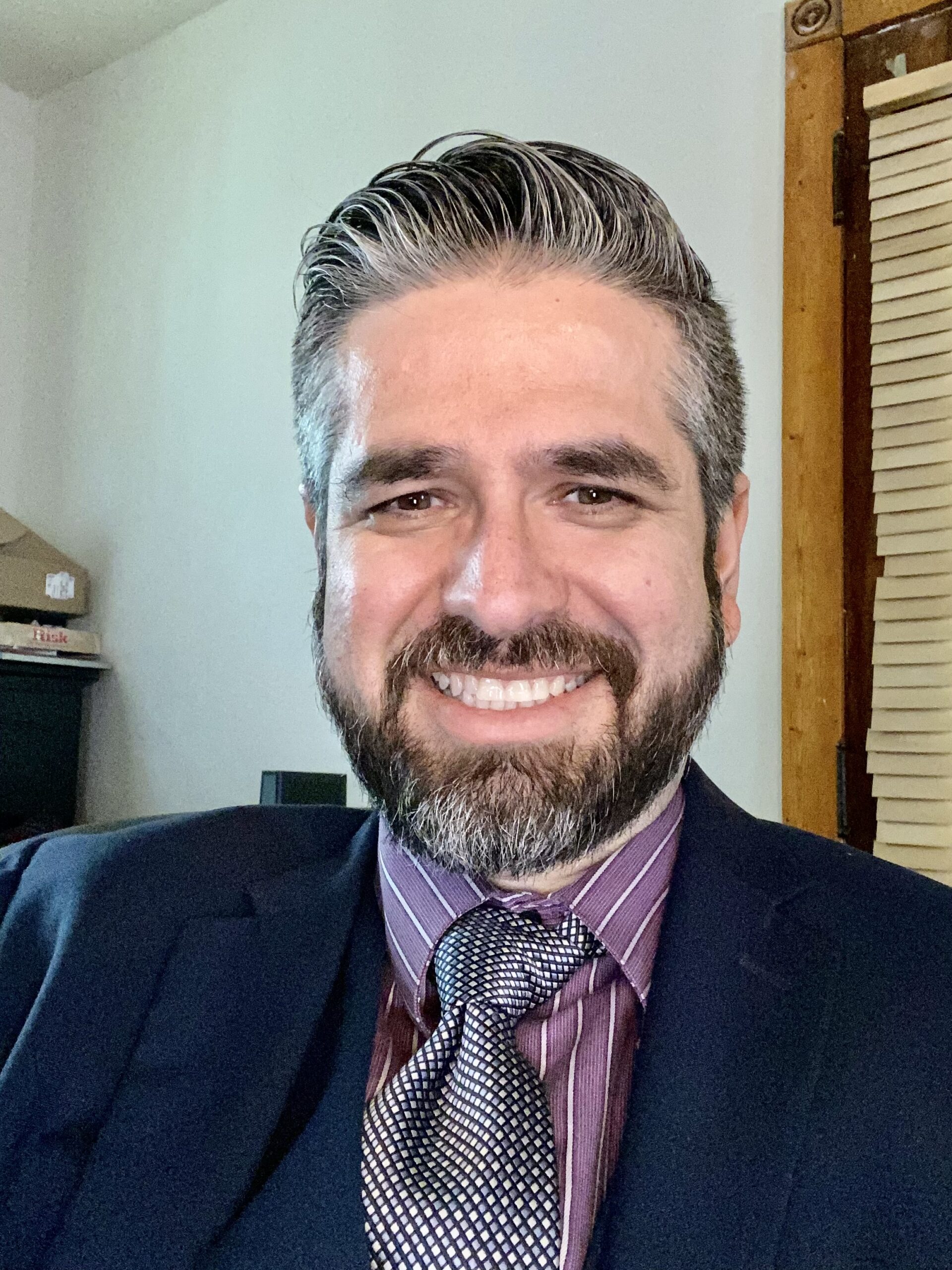Smiling man in formal attire with beard and tie, sitting indoors near window blinds.