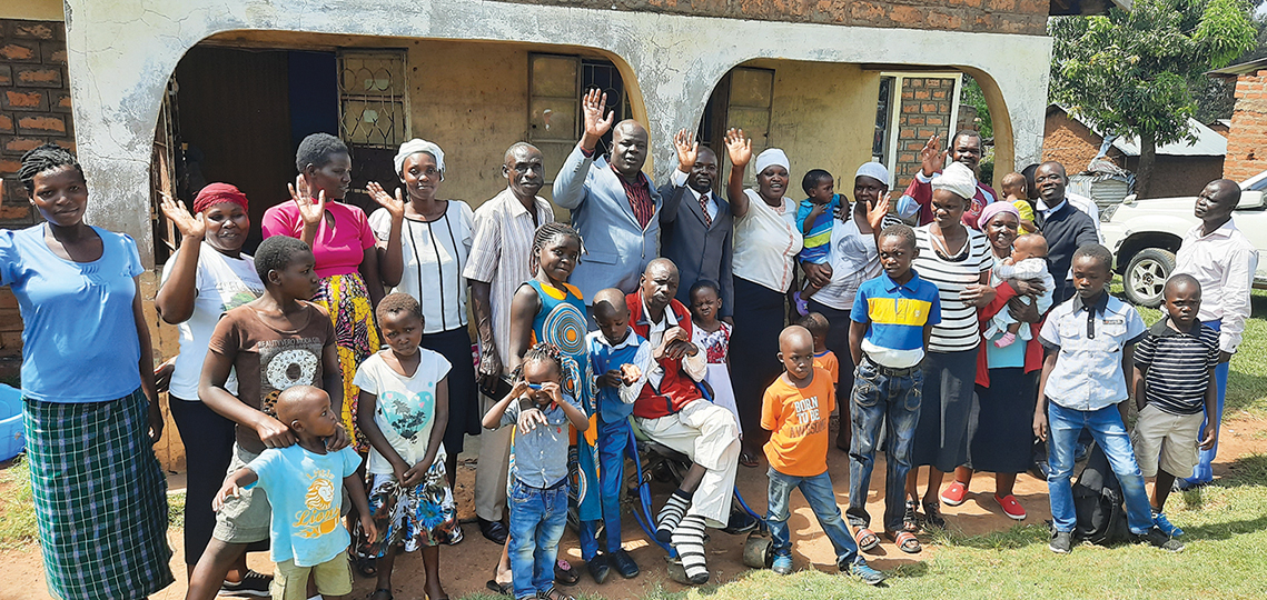 Group of people smiling and waving outside a house, enjoying a sunny day in a rural setting.
