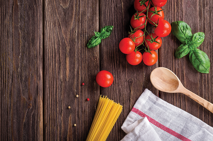 Fresh ingredients on wooden table: cherry tomatoes, basil, spaghetti, peppercorns, wooden spoon, and kitchen towel.