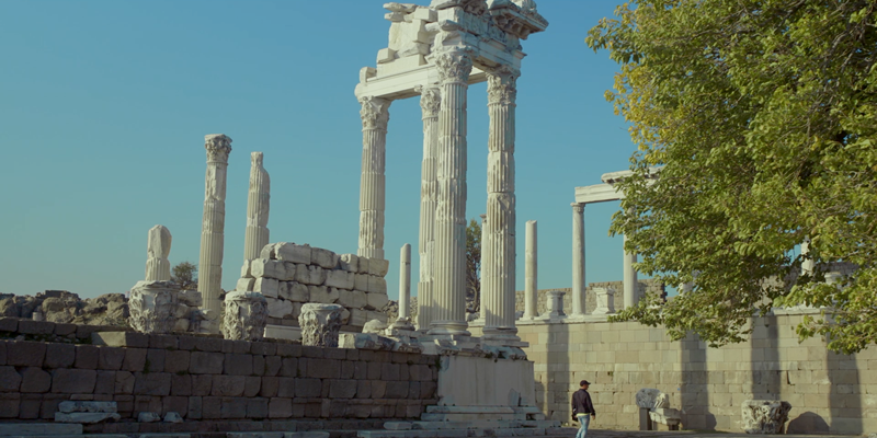 Ancient Greek ruins with tall columns under a clear blue sky, surrounded by trees and a person exploring the site.