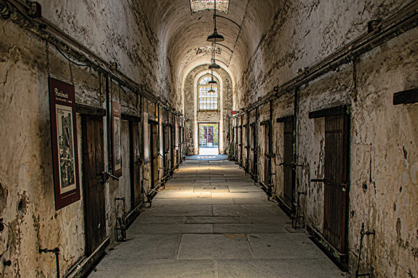 Abandoned prison corridor with peeling walls and old cells, leading to a sunlit exit.