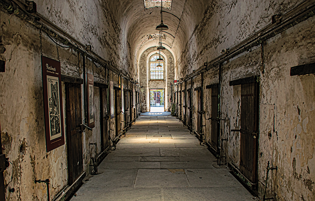 Abandoned prison corridor with peeling walls and old cells, leading to a sunlit exit.