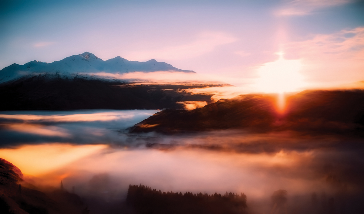 Mountain landscape at sunrise with mist and soft pastel colors, highlighting snowy peaks and a vibrant sky.