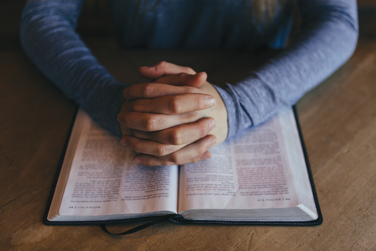 Hands folded in prayer on an open Bible, symbolizing faith and reflection.