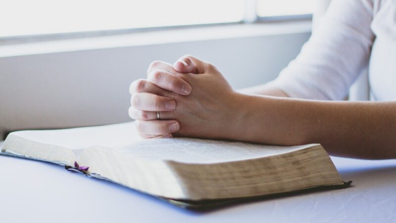 Person praying with hands clasped over an open Bible by a window.