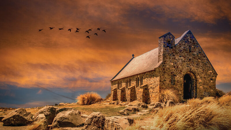 Stone church under vibrant orange sky with flying birds, surrounded by rocks and grass.