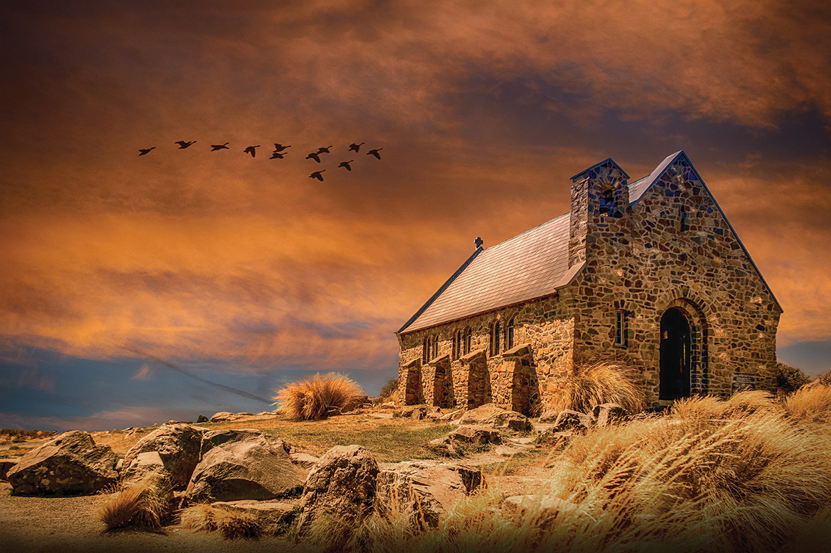 Stone church under vibrant orange sky with flying birds, surrounded by rocks and grass.