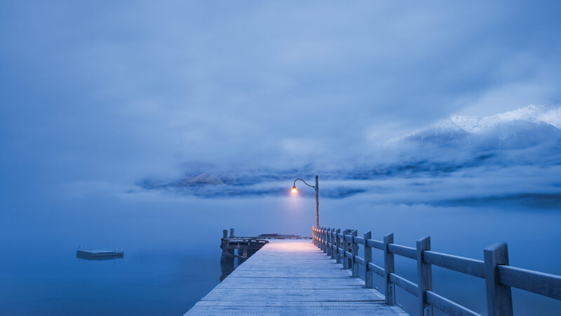 Moody pier and lamp post over calm lake, enveloped in fog, with mountains in the background at dawn.