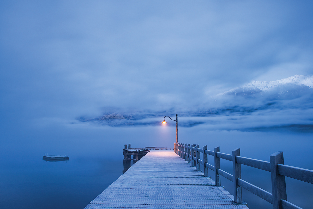 Moody pier and lamp post over calm lake, enveloped in fog, with mountains in the background at dawn.