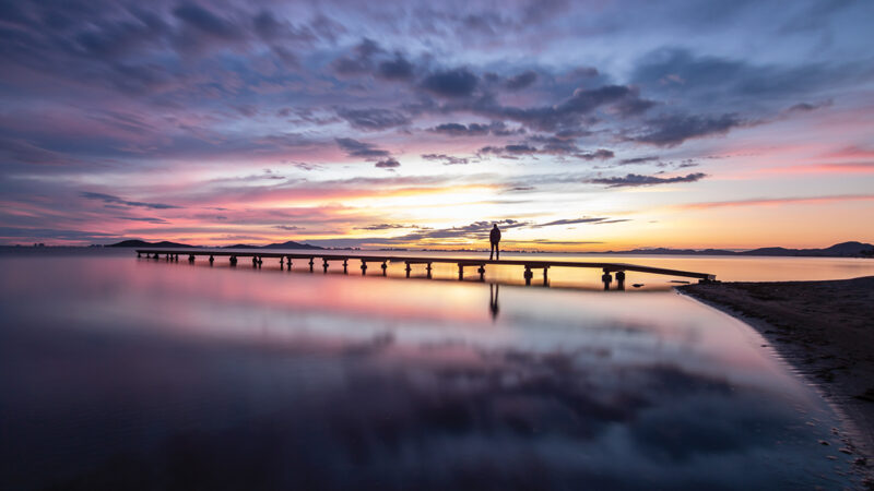 Serene sunset over a calm lake with a silhouette standing on a long pier.