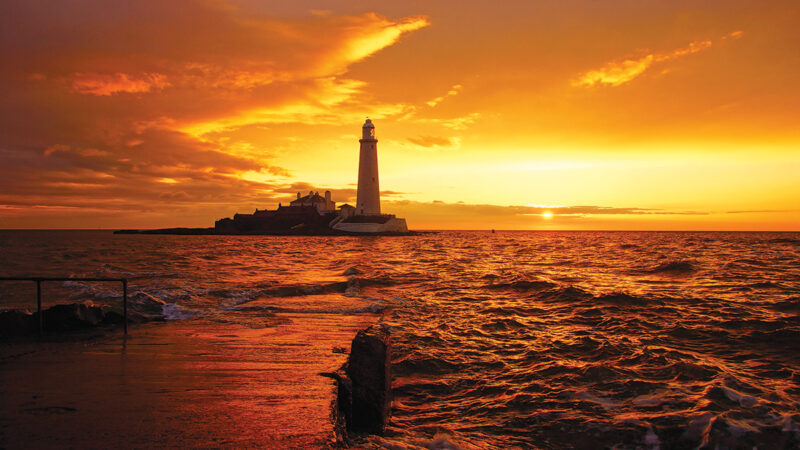 Golden sunset over the sea with a silhouette of a lighthouse on the horizon.