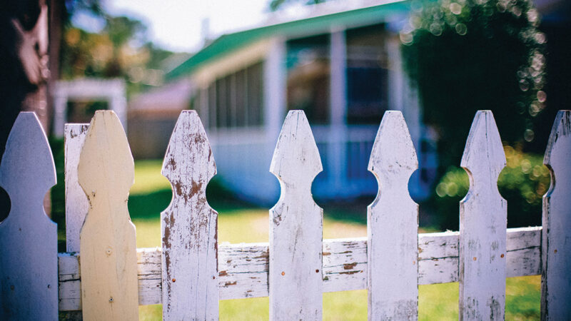 Weathered white picket fence with blurred house background, summer day ambiance.