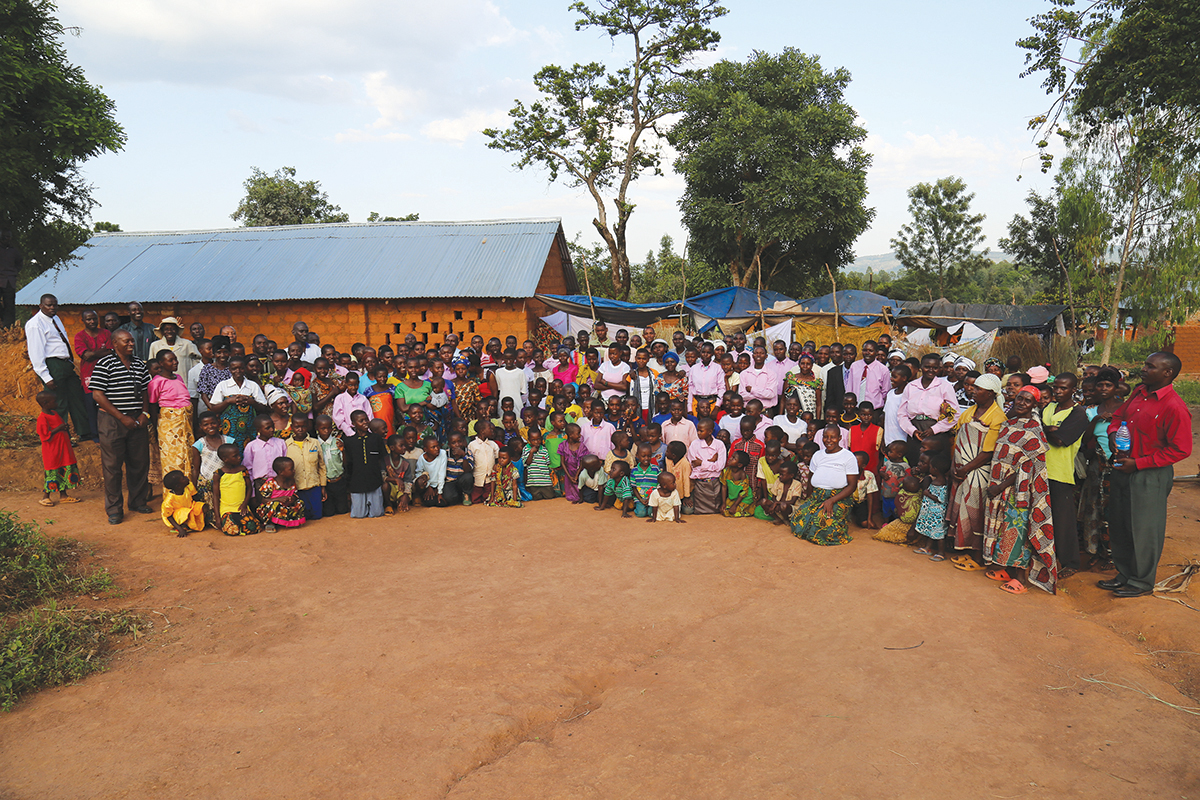 Large group of people gathered outside a community building surrounded by trees and a clear sky.