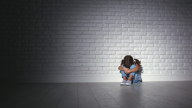 Lonely child sitting against a white brick wall, hugging knees, conveying sadness and isolation in a dimly lit room.