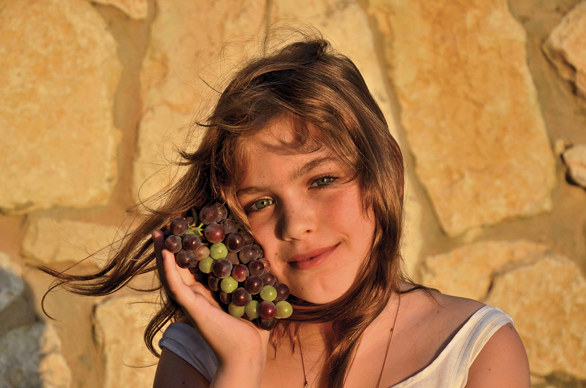 Girl smiling with grapes against stone wall in warm sunlight.