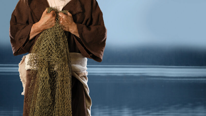 Person in brown robe holds a fishing net by a calm lake backdrop, conveying a historical or biblical theme.