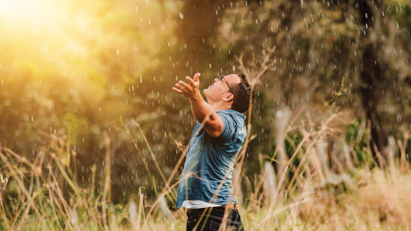 Man joyfully embracing rain in sunlit field, arms outstretched, surrounded by grass and trees.