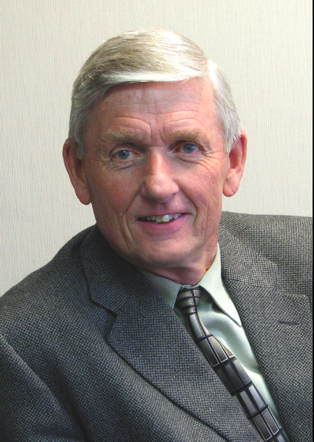 Elderly man in a suit smiling, with gray hair and blue eyes, against a neutral background.