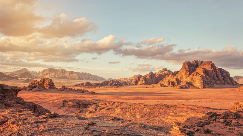 Stunning desert landscape with rocky mountains, golden sand, and cloudy sky at sunset.