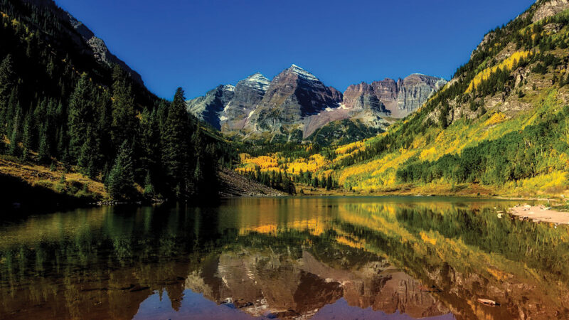 Scenic mountain landscape with autumn colors reflected in a calm lake under a clear blue sky.