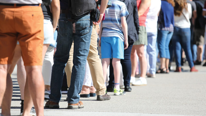 A diverse group of people standing in line outdoors on a sunny day, showing casual summer clothing and footwear.