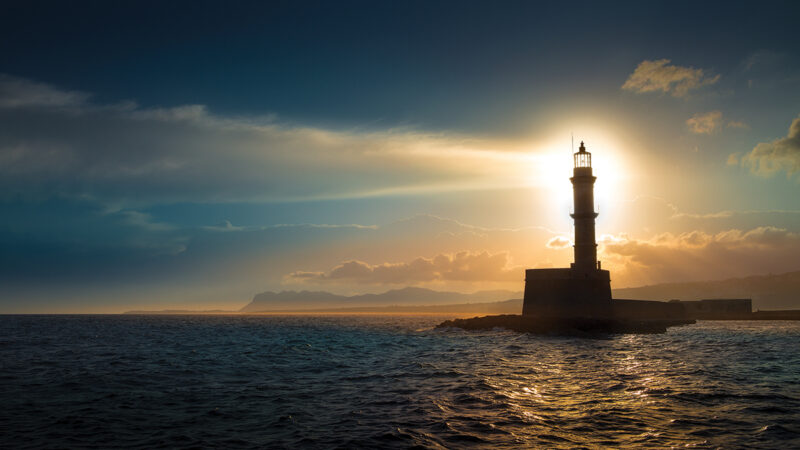 Lighthouse at sunset over ocean waves, casting a silhouette against dramatic clouds and distant mountains.