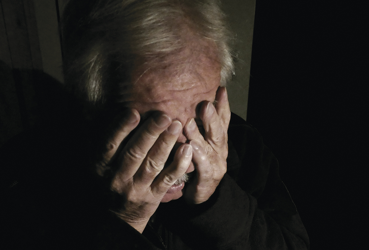 Elderly person covering face with hands in dim lighting, conveying emotion and vulnerability.