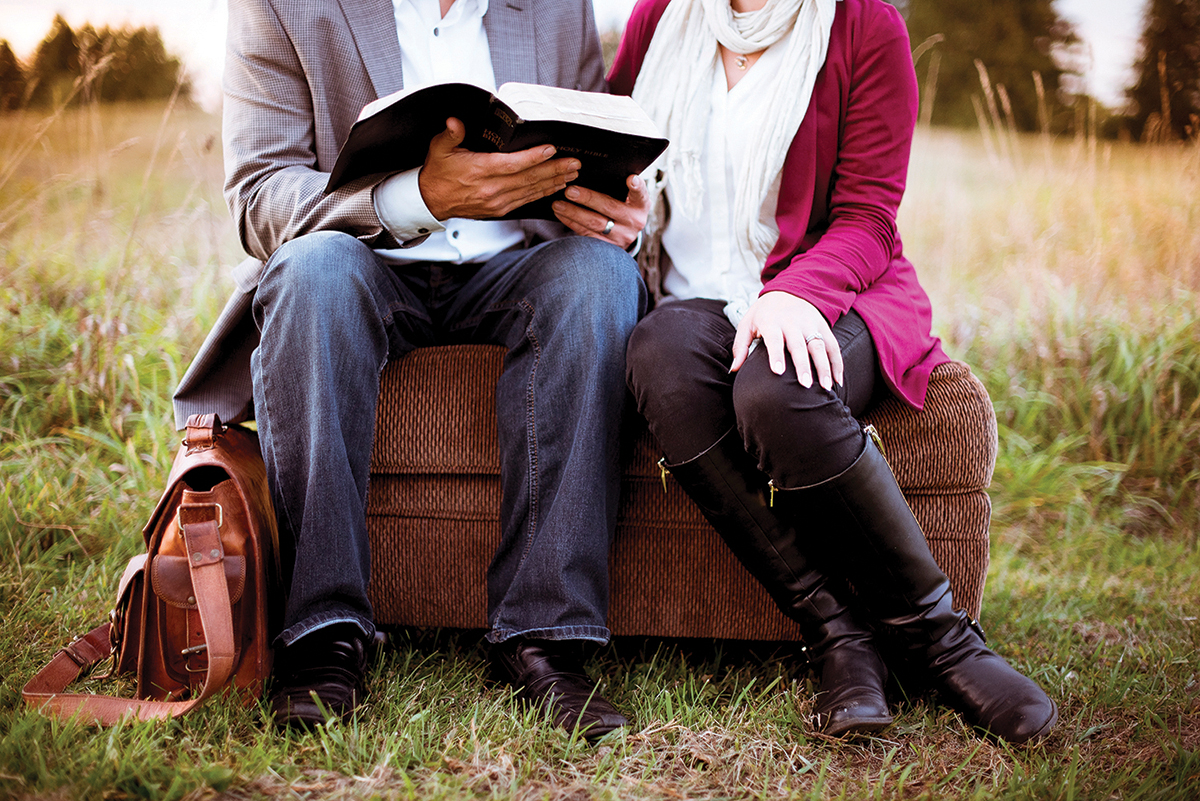 Couple reading a book together on a couch in a field, with a brown leather bag beside them.